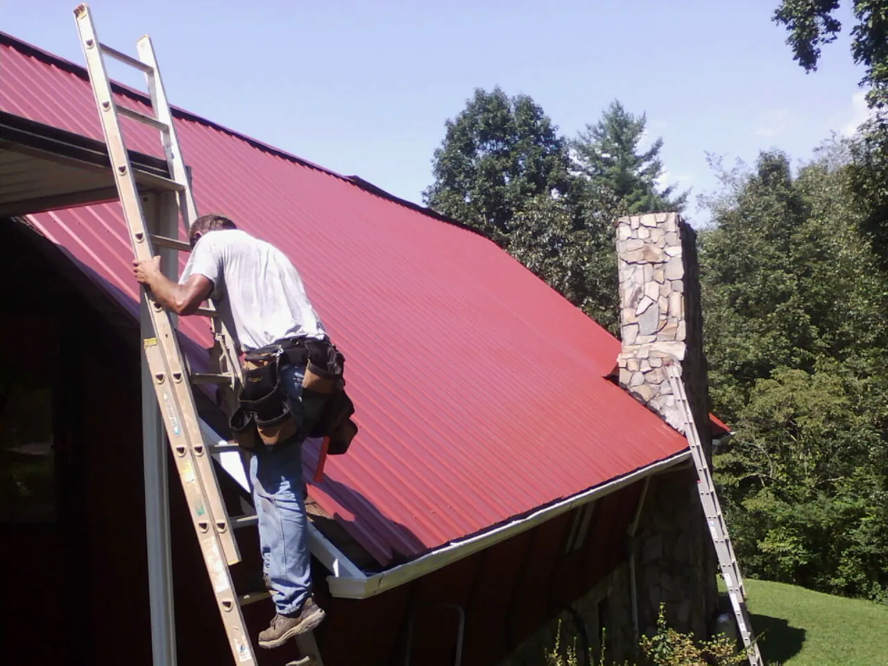 Roofer on a ladder beside a red metal roof and stone chimney — Soffit & Fascia Repair by Roofing Brothers in Rootstown